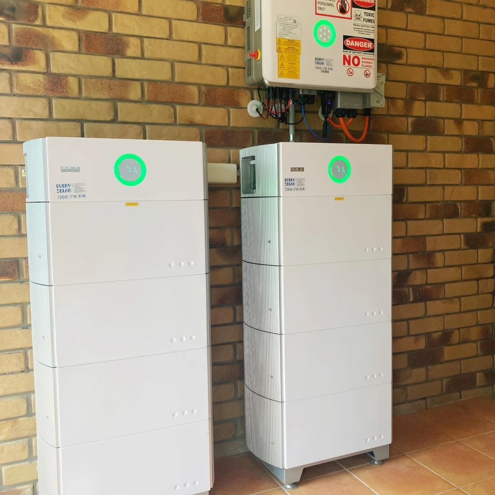 Two tall white SAJ battery units with green ring displays installed side-by-side against a brick wall, with electrical equipment above. Two tall white SAJ battery units with green ring displays installed side-by-side against a brick wall, with electrical equipment above.